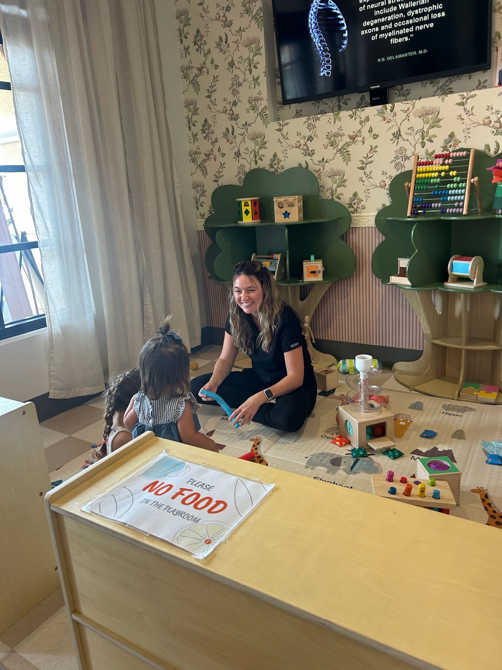Carly plays on the floor with two kids in the Little Roots play area.