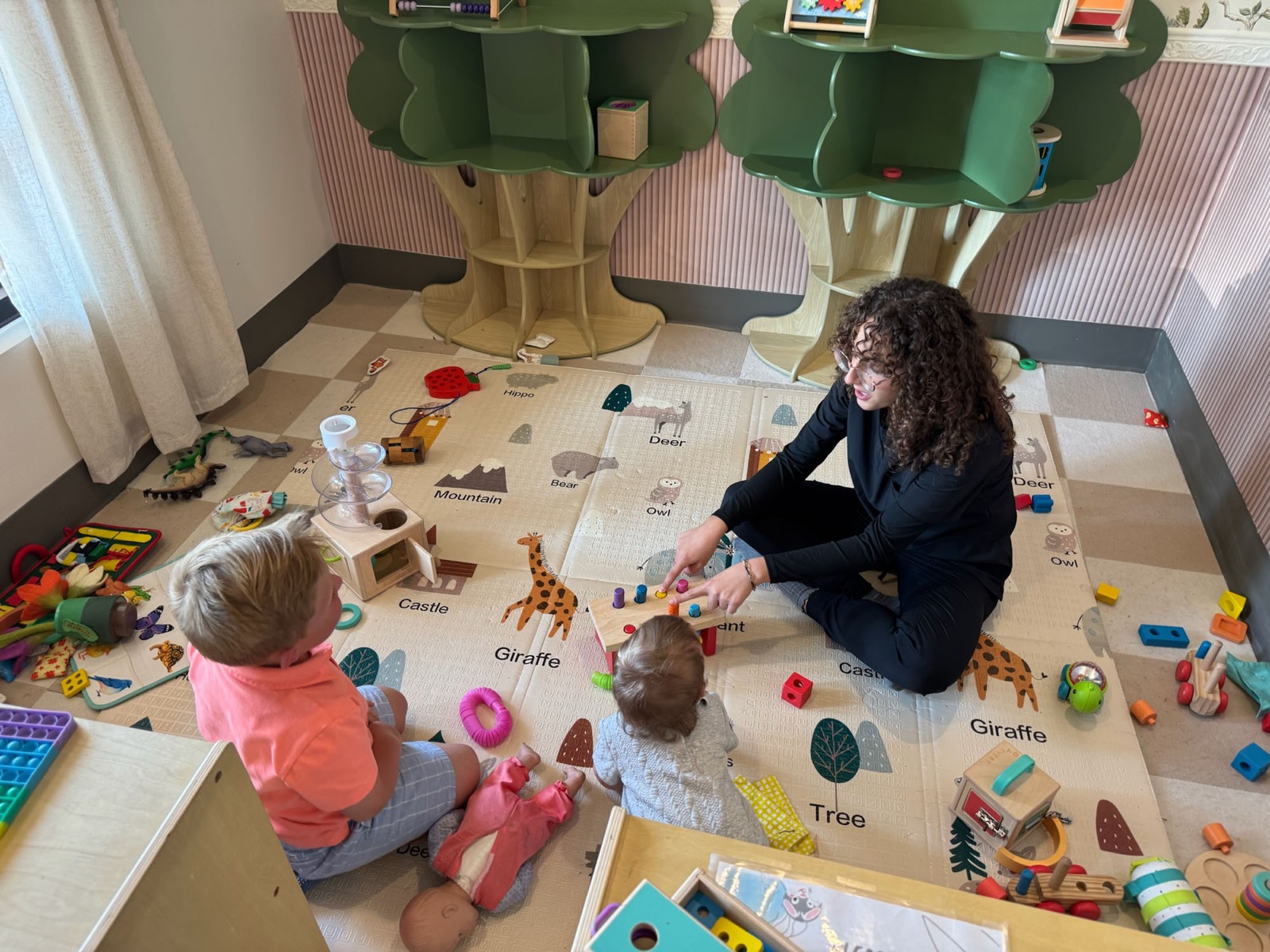 Yohanna plays on the floor with two kids in the Little Roots play area.
